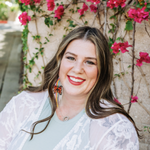 Rachel is wearing a white lace shawl, over a green shirt, and is smiling. She is in front of bright pink flowers and has a butterfly and rainbow earring set on.